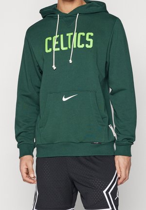 Homme portant un sweat à capuche vert des Celtics avec des cordons blancs et un short de sport noir avec des motifs géométriques blancs.