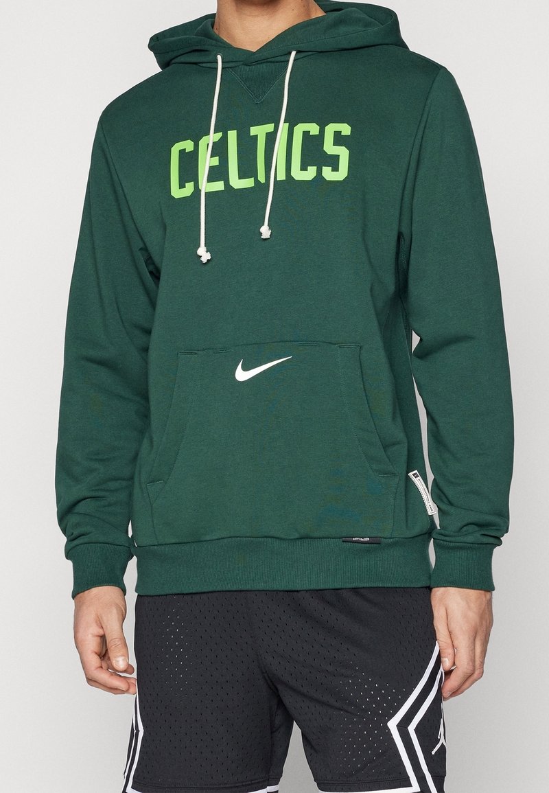 Homme portant un sweat à capuche vert des Celtics avec des cordons blancs et un short de sport noir avec des motifs géométriques blancs.