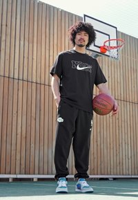 Černé tričko Nike s logem, černé tepláky s logem a modré tenisky. Držící basketbal na hřišti s dřevěným pozadím.