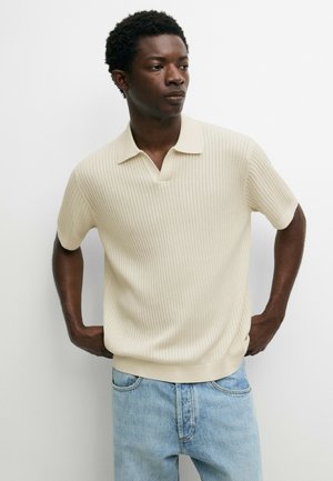 Junger Mann mit kurzen lockigen Haaren, der ein cremefarbenes, geripptes, kurzärmeliges Poloshirt und hellblaue Jeans trägt, steht mit den Händen in der Hüfte.