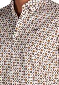 Witte button-up shirt met een herhaald bloem- en sterpatroon in oranje, bruin en groen. Inclusief een klein logo op de borst.