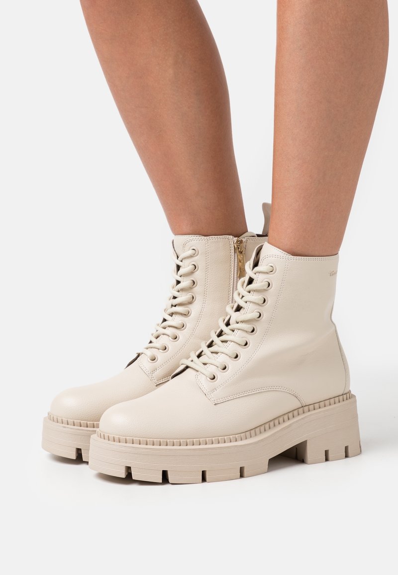 Bottines beiges à lacets avec semelles épaisses et rainurées, fermetures éclair latérales, portées sur des jambes nues sur un fond uni.