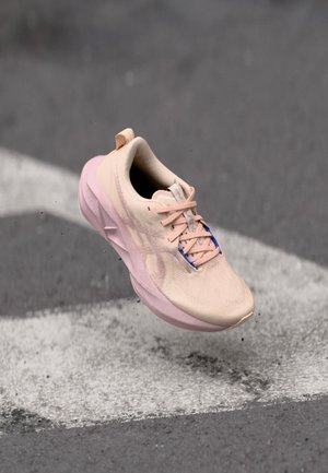 Baskets de sport rose clair avec un dessus texturé, semelle rembourrée et lacets bleus contrastants, montrées en l'air au-dessus d'une surface texturée.