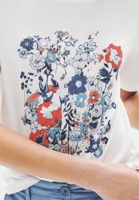 Camiseta blanca de manga corta con un estampado floral en rojo, azul y blanco. El diseño incluye varias formas de flores y detalles intrincados.
