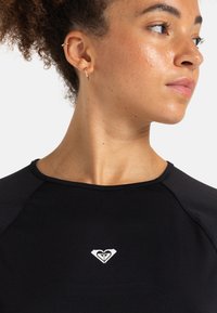 Nainen, jolla on kiharat hiukset ja pienet kultaketjut, katsoo oikealle, yllään musta paita, jossa on edessä pieni valkoinen sydänlogo.