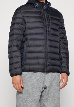 Mężczyzna w czarnej pikowanej kurtce z kapturem, z kieszenią na piersi zapinaną na zamek, oraz w szarych spodniach dresowych, widoczny od ramion do bioder.