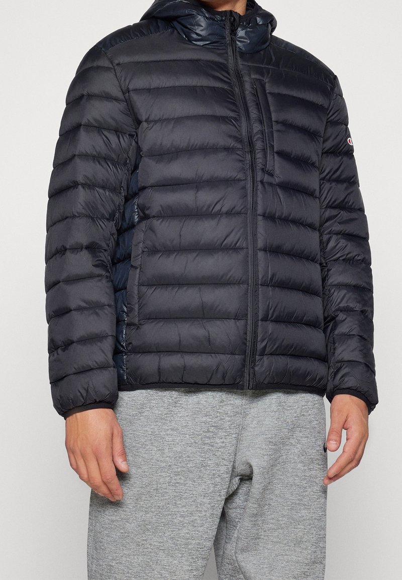 Homme portant une veste matelassée noire à capuche avec une poche poitrine zippée et un pantalon de survêtement gris, visible des épaules jusqu'aux hanches.
