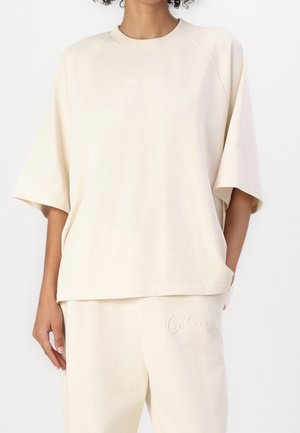 Person trägt locker sitzendes off-white kurzärmeliges Oberteil und passende Hose mit subtil gesticktem Branding auf Brust und Oberschenkel.
