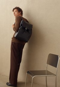Mochila de cuero negro con solapa y detalle de hebilla, llevada por un modelo que viste un suéter de lana marrón y pantalones oscuros, al lado de una silla de metal.