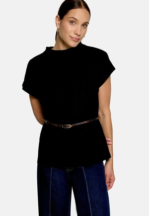 Femme aux cheveux bruns portant un haut noir à manches courtes ceinturé à la taille, avec un jean bleu, debout devant un fond clair uni.