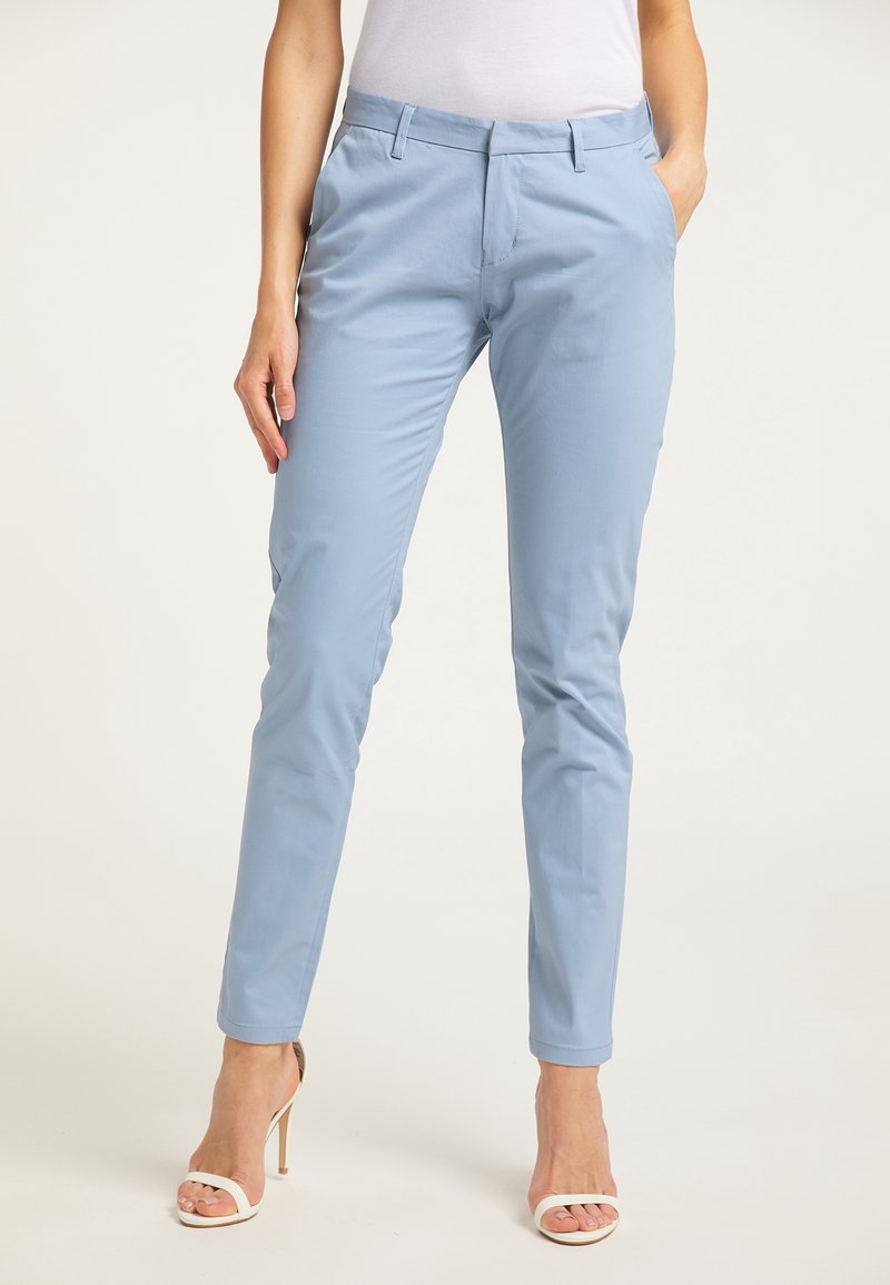 Femme portant un pantalon slim bleu clair et des sandales blanches à talons hauts, debout avec une main dans la poche, sur un fond uni.