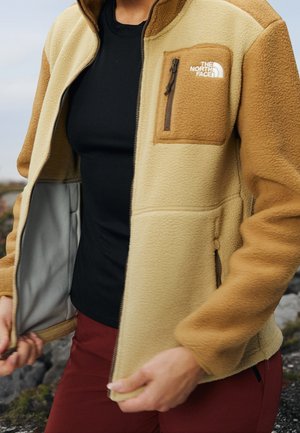 Osoba nosząca beżowo-brązową kurtkę polarową z kieszenią na zamek na piersi oraz logo "The North Face", rozpina kurtkę na zewnątrz.