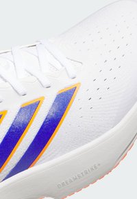 Λευκό αθλητικό παπούτσι με υφή στην επάνω επιφάνεια, μοβ και πορτοκαλί λεπτομέρειες, και οπές για αερισμό. Η σόλα φέρει το σήμα "DREAMSTRIKE+".