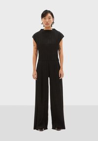 Zwarte geribde jumpsuit met een hoge halslijn, kapmouwen en wijde benen, gepresenteerd op een model tegen een effen achtergrond.