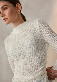 White textured top with long sleeves, featuring a high neckline and subtle gathering details at the waist, made from a lightweight fabric.