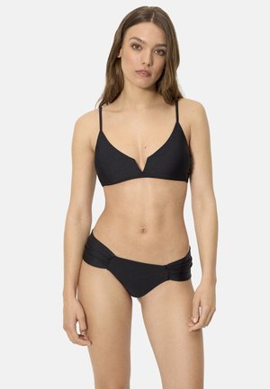Mujer con cabello largo y castaño, vistiendo un bikini negro de dos piezas con un top en forma de V con muesca y pantalones con fruncido lateral, posando frente a un fondo blanco liso.