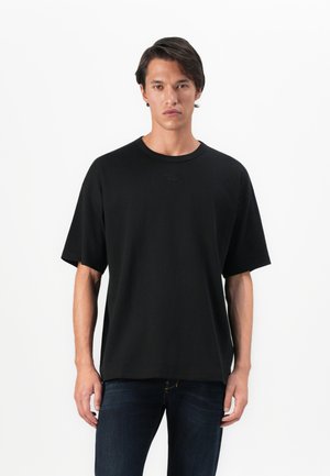 Jeune homme aux cheveux bruns portant un t-shirt noir oversize uni et un jean bleu foncé, se tenant devant un fond blanc.