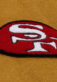 Logo ricamato rosso e bianco su materiale in feltro con sfondo marrone, caratterizzato da un design testurizzato e contorno nero.