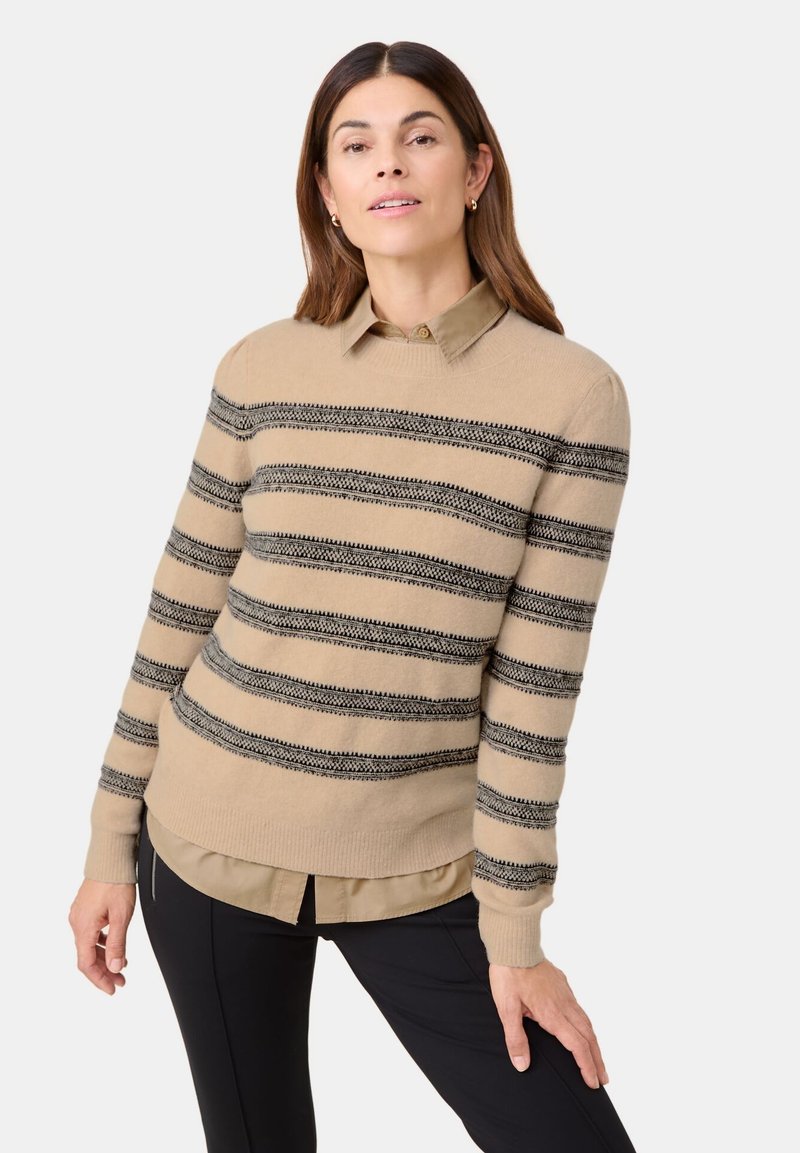 Beige sweater with black stripes, round neckline, and ribbed cuffs, layered over a collared shirt, worn with black trousers.