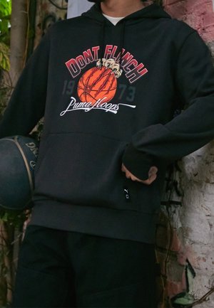 Personne portant un sweat à capuche noir avec un graphique de basketball, des griffures et le texte "DONT FLINCH Puma Hoops", tenant un ballon de basketball noir.