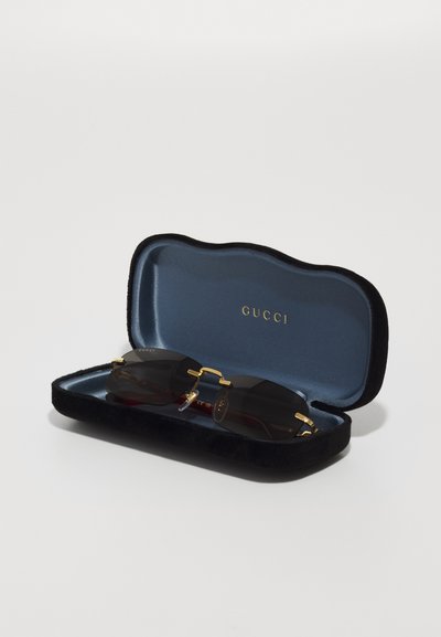 Gafas de sol Gucci en un estuche de terciopelo negro, con un interior azul. Las lentes son oscuras con acentos dorados y un diseño de puente único.