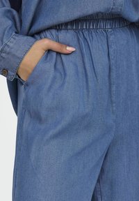 Hand resting in pocket of loose elastic-waist denim pants worn with matching denim long-sleeve shirt.