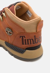 Zapatilla de cuero marrón con parte superior en tono marrón claro, acentos texturizados, ojales de metal y suela de goma negra. Cuenta con el logotipo "Timberland" en relieve.