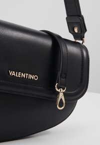 Borsa a spalla in pelle nera con patta piegata, hardware dorato e logo "VALENTINO". Texture liscia con una silhouette leggermente curvata.