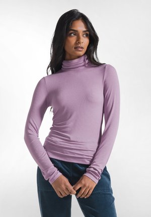 Mujer con cabello largo y oscuro, vestida con un jersey de cuello alto ajustado color lavanda y pantalones azul oscuro, de pie con las manos ligeramente entrelazadas a la altura de la cintura.