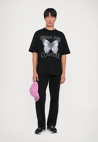 Μαύρη oversized μπλούζα με γραφικό πεταλούδας και κείμενο "Versace Jeans Couture", σε συνδυασμό με μαύρο παντελόνι και ροζ καπέλο.