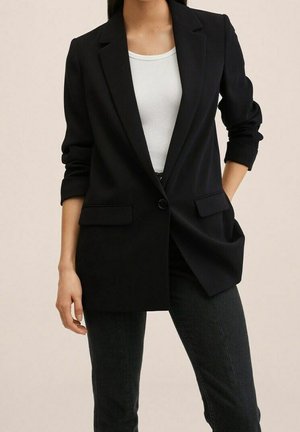 Femme portant un blazer noir à un seul bouton sur un débardeur blanc côtelé avec un pantalon foncé ajusté, une main dans la poche.