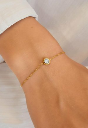Delicate gold chain bracelet with a single round clear gemstone centerpiece worn on a wrist against a light fabric background.