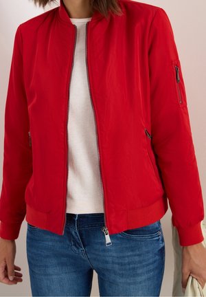 Frau trägt eine leuchtend rote Bomberjacke mit Reißverschluss über einem weißen Pullover und blauen Jeans, hält eine hellfarbige Tasche in der rechten Hand.