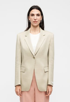 Femme aux cheveux raides et foncés porte un blazer beige sur un top blanc et un pantalon rose pâle, debout devant un fond clair uni.