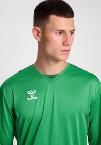 Groen sporttruitje met een V-hals ontwerp, gemaakt van een gladde stof. Kenmerkt zwarte accenten op de schouders en een wit logo op de borst.