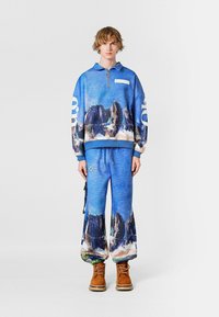 Sudadera azul con cremallera y estampado de montaña, combinada con pantalones de corte holgado que presentan el mismo diseño. Botas marrones completan el atuendo.