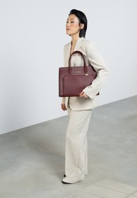 Burgundy leather tote bag with two short handles, a zip closure, and front pocket, paired with a light-colored suit and black shoes.