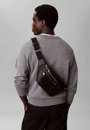 Uomo con maglione grigio che indossa una borsa a tracolla in pelle nera con cerniera, visto da dietro su uno sfondo grigio uniforme.