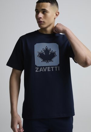 Mladý muž upravující si tmavě modré tričko s puntíkovaným motivem javorového listu a nápisem "ZAVETTI" na neutrálním šedém pozadí.