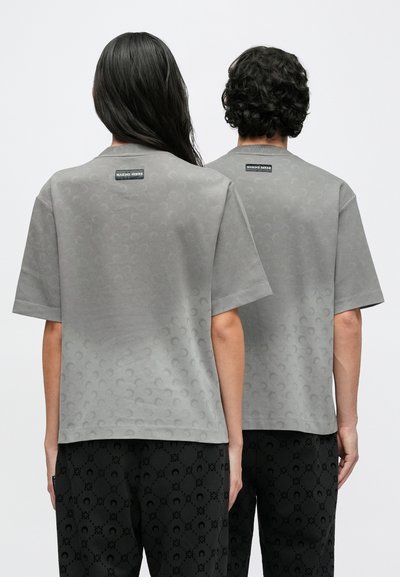 Szare t-shirty z krótkim rękawem z subtelnym okrągłym wzorem i metką marki na plecach. Noszone z czarnymi spodniami w wzory.