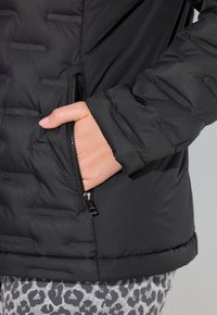 Černá bundy s péřovou izolací, horizontálně prošívaná, s kapsou na zip a žebrovanými manžetami. Texturovaná tkanina s hladkým povrchem.