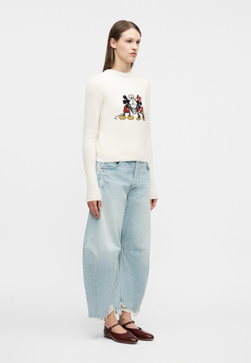 Frau trägt einen weißen Pullover mit Mickey- und Minnie-Design, hellblaue Jeans mit weitem Bein und ausgefranstem Saum sowie dunkelrote Mary Jane Schuhe.