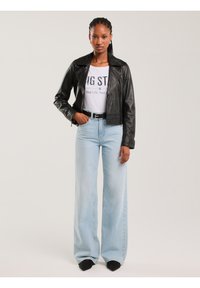 Naine punutud patsidega, musta nahktagi, valge graafilise t-särgi, hele siniste laia jalaga teksade ja mustade teravate kingadega seismas.