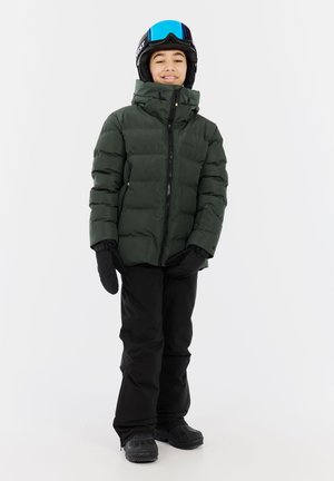 Grüne Daunenjacke mit hohem Kragen, schwarze Schneehose und schwarze Fäustlinge. Schwarze Skischuhe. Blaugraue Skibrille auf dem Kopf.