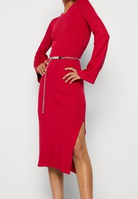 Rotes, geripptes Strickkleid mit langen Glockenärmeln, seitlichem Schlitz und silberner Kettengürtelschließe, die die Taille betont. Glatte Textur und figurbetonter Schnitt.