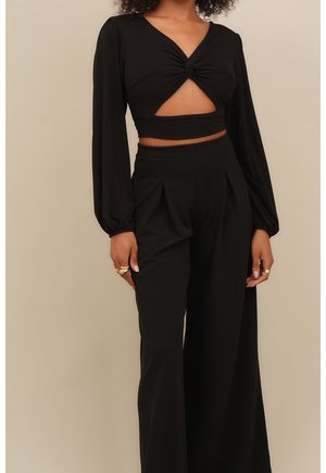 Nainen pukeutuneena mustaan pitkähihainen lyhyt toppiin, jossa etukappaleen leikkaus, ja korkeavyötäröisiin leveälahkeisiin mustiin housuihin, beige tausta.