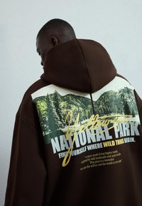 Hnědá mikina s kapucí, která má grafiku Národního parku Yellowstone, se zelenými stromy a bílým textem. Kapuce je přišita; látka vypadá měkce.