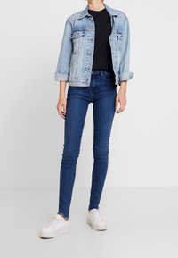 Světle modrá denimová bunda s klasickým designem, kombinovaná s černým tričkem a tmavě modrými skinny džíny. Bílá teniska doplňuje celý outfit.