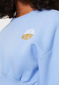Ljusblå sweatshirt med "GOOD TIMES" broderat i vitt och guld. Mjuk tyg med avslappnad passform och sänkt axeldesign.