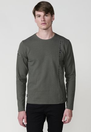 Camiseta de manga larga verde oliva con textura acanalada, cuello redondo y detalle de texto negro vertical en el lado izquierdo; combinar con pantalones negros.